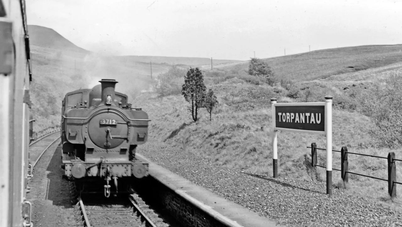 Torpantau Station: meeting a Brecon - Newport train.
View northward, towards Torpantau Tunnel, Talybont and Brecon: ex-Brecon & Merthyr (Brecon - Dowlais section), later GWR. The down train, headed by standard '8750' class 0-6-0PT No. 3712 (built 11/36) has just surmounted the fearsome climb of seven miles (the "Seven-Mile Bank") at about 1-in-40 from Talybont-on-Usk, while my train headed for Brecon has come up a similar climb from Pontsticill Junction. The narrow-gauge Brecon Mountain Railway from Pant terminates just short of here but aims to reach this point. On the left is the outline of the Brecon Beacons.